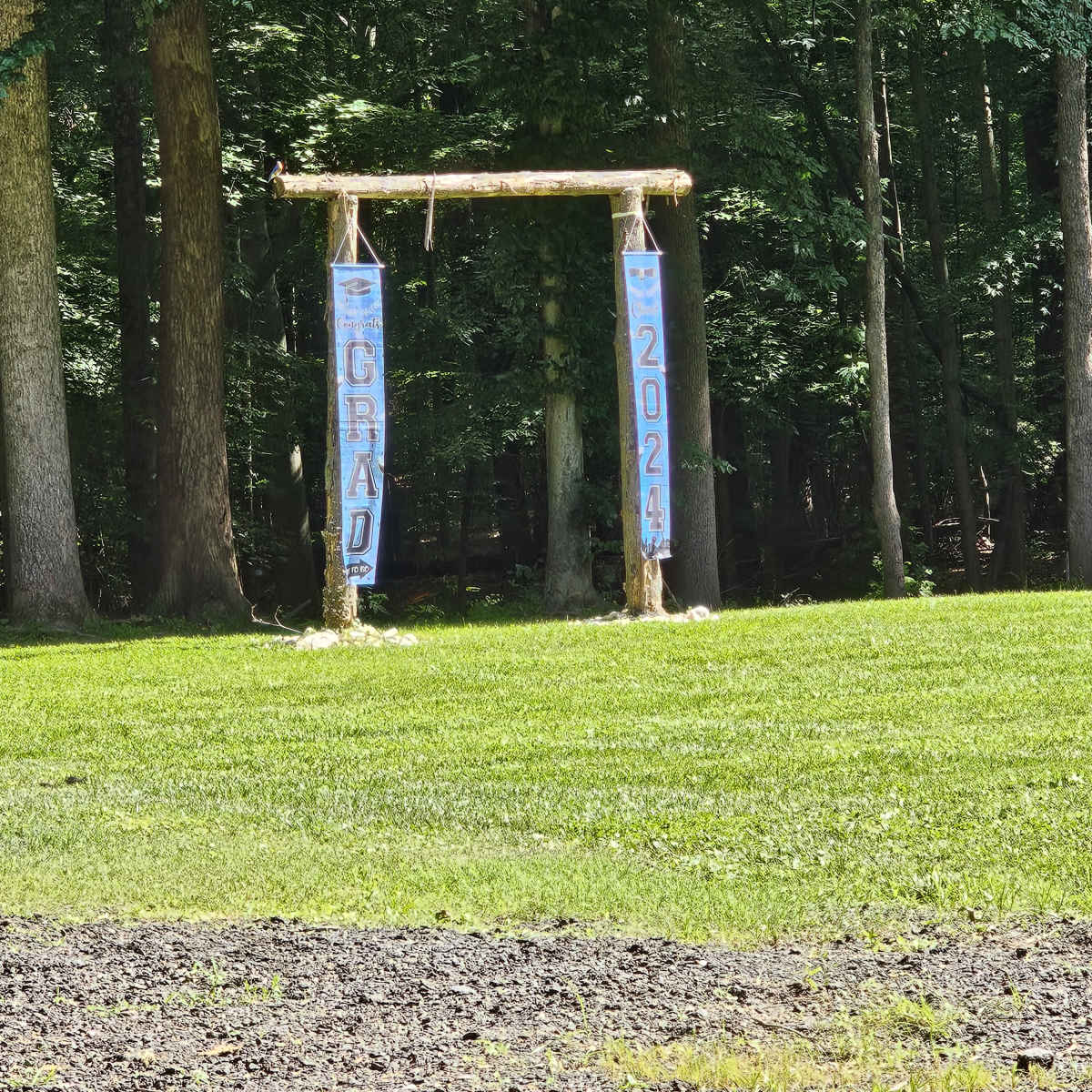 A wooden archway displayed with banners reading "GRAD" and "2024" stands on a grassy area surrounded by trees.