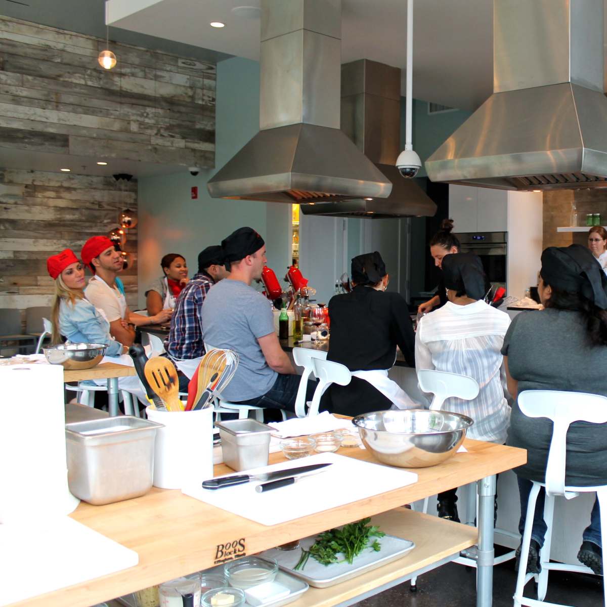 A group of people wearing red chef hats sits around a kitchen island, observing a chef giving instructions in a modern cooking classroom.