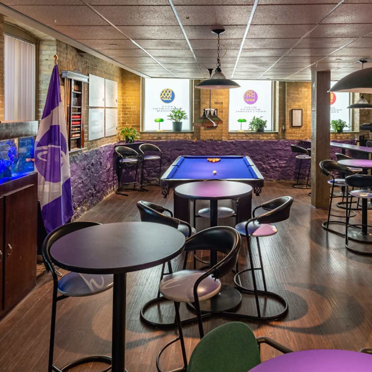 A spacious game room with a billiards table, tables and chairs, an aquarium, and vending machines against a backdrop of exposed brick walls.