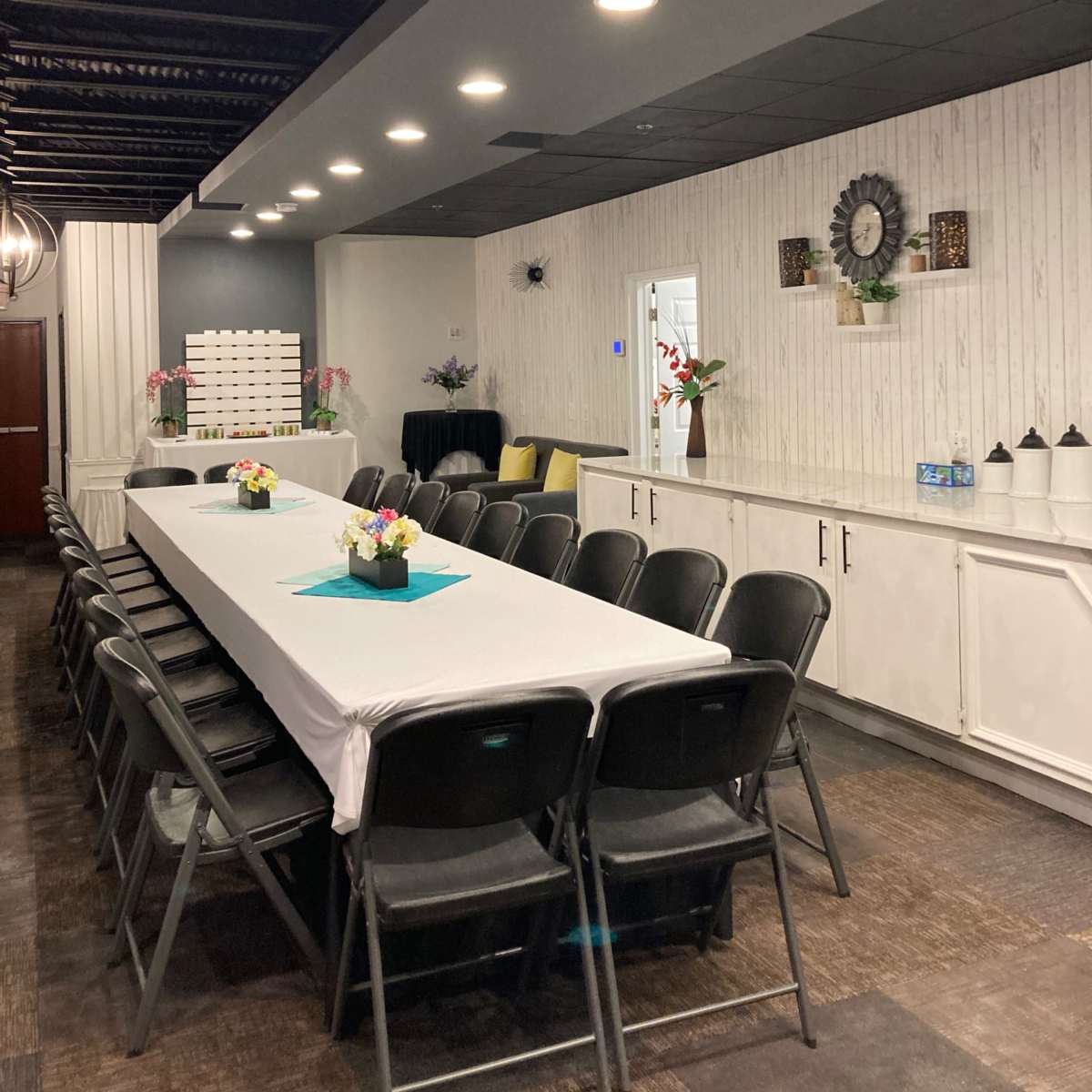 A well-lit meeting room with a long white table surrounded by black chairs, decorated with flowers and featuring a clock on the wall.