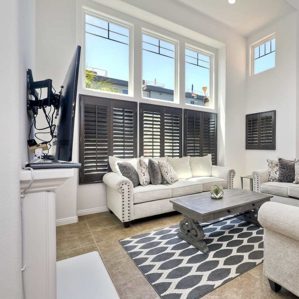 A spacious living room with large windows, light-colored furniture, and a coffee table on a patterned rug.
