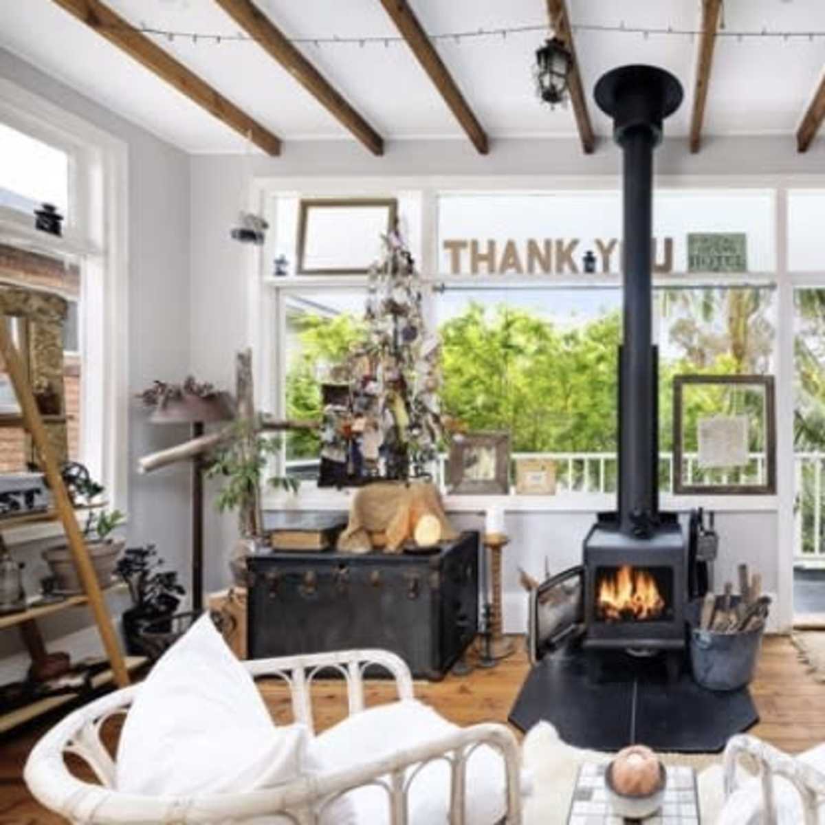 A cozy living room features a central black wood stove surrounded by light-colored furniture, large windows showcasing greenery, and wooden beams on the ceiling.