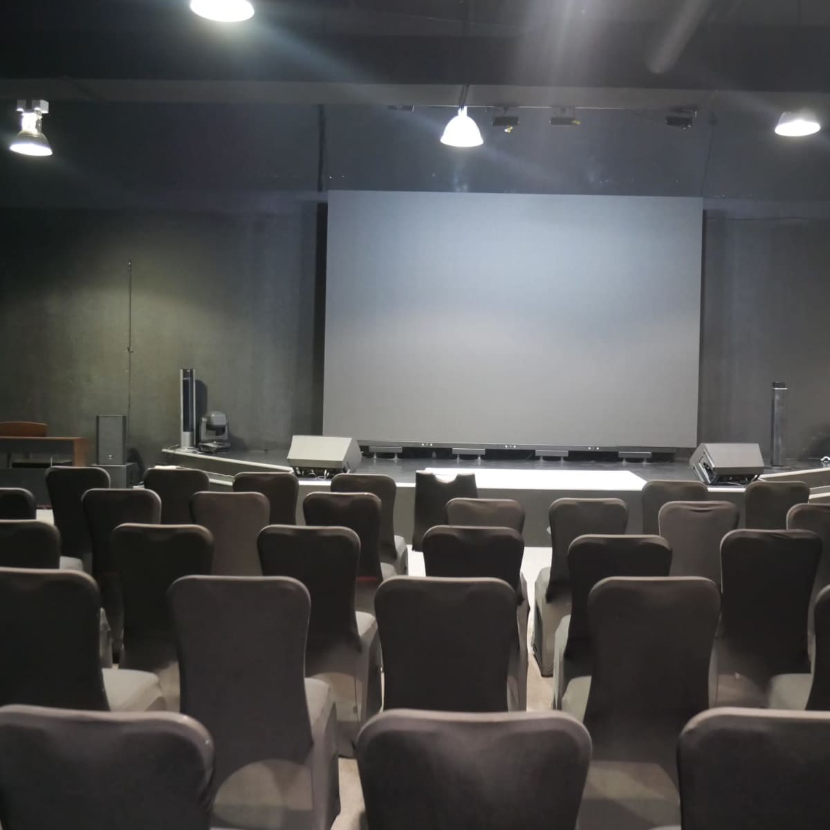 A row of empty chairs faces a large blank screen on a darkened stage in a modern auditorium.