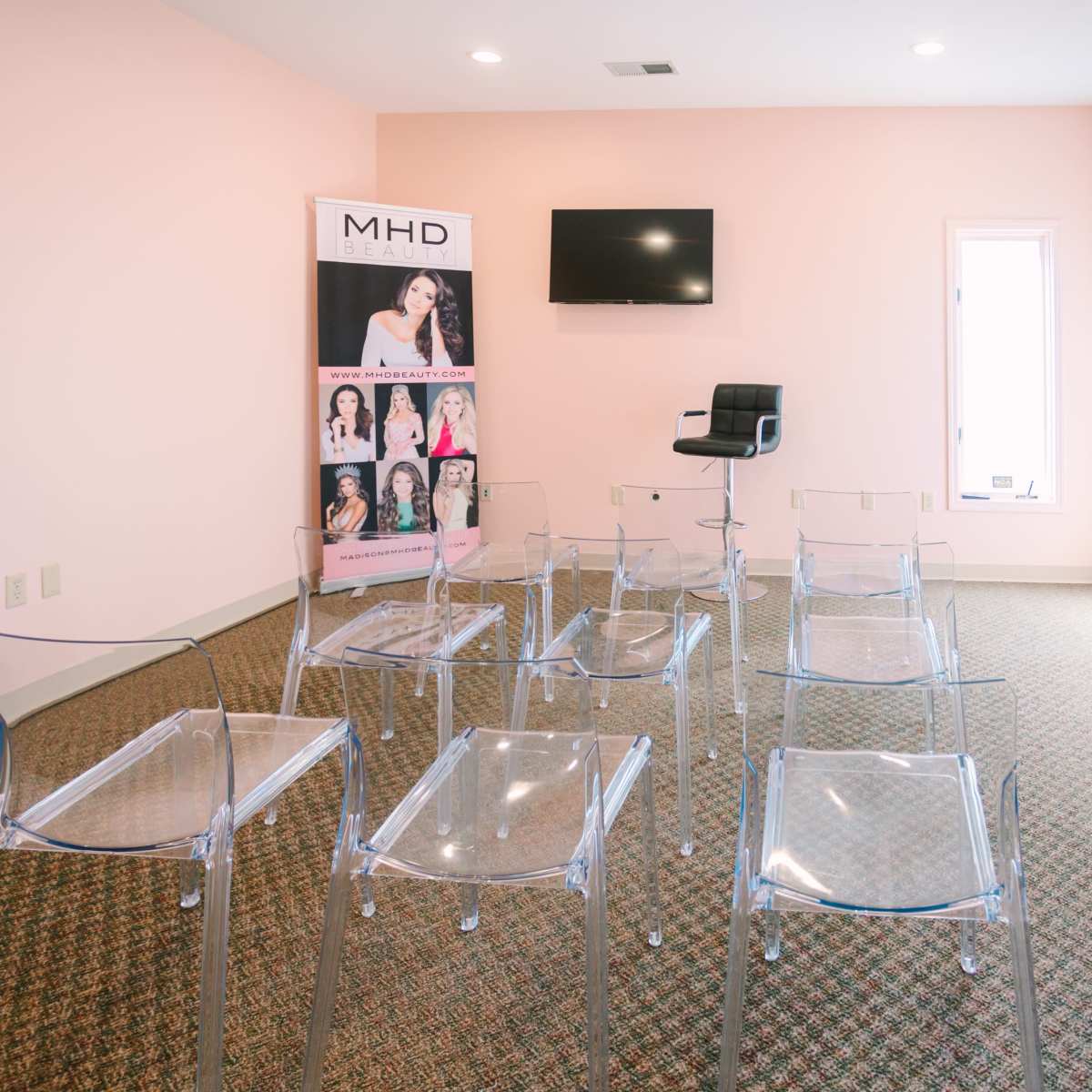 A room with a light pink wall, featuring a row of clear chairs, a black stool, a television on the wall, and a banner displaying images of individuals.