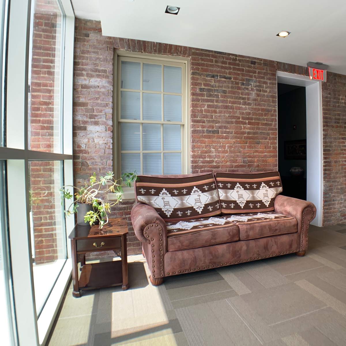 A brown sofa with a patterned throw placed against a brick wall next to a small table and a large window.