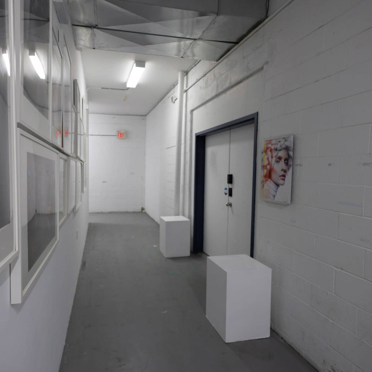 A narrow hallway with white walls features framed photographs on one side and a door with a small portrait hanging adjacent to it.