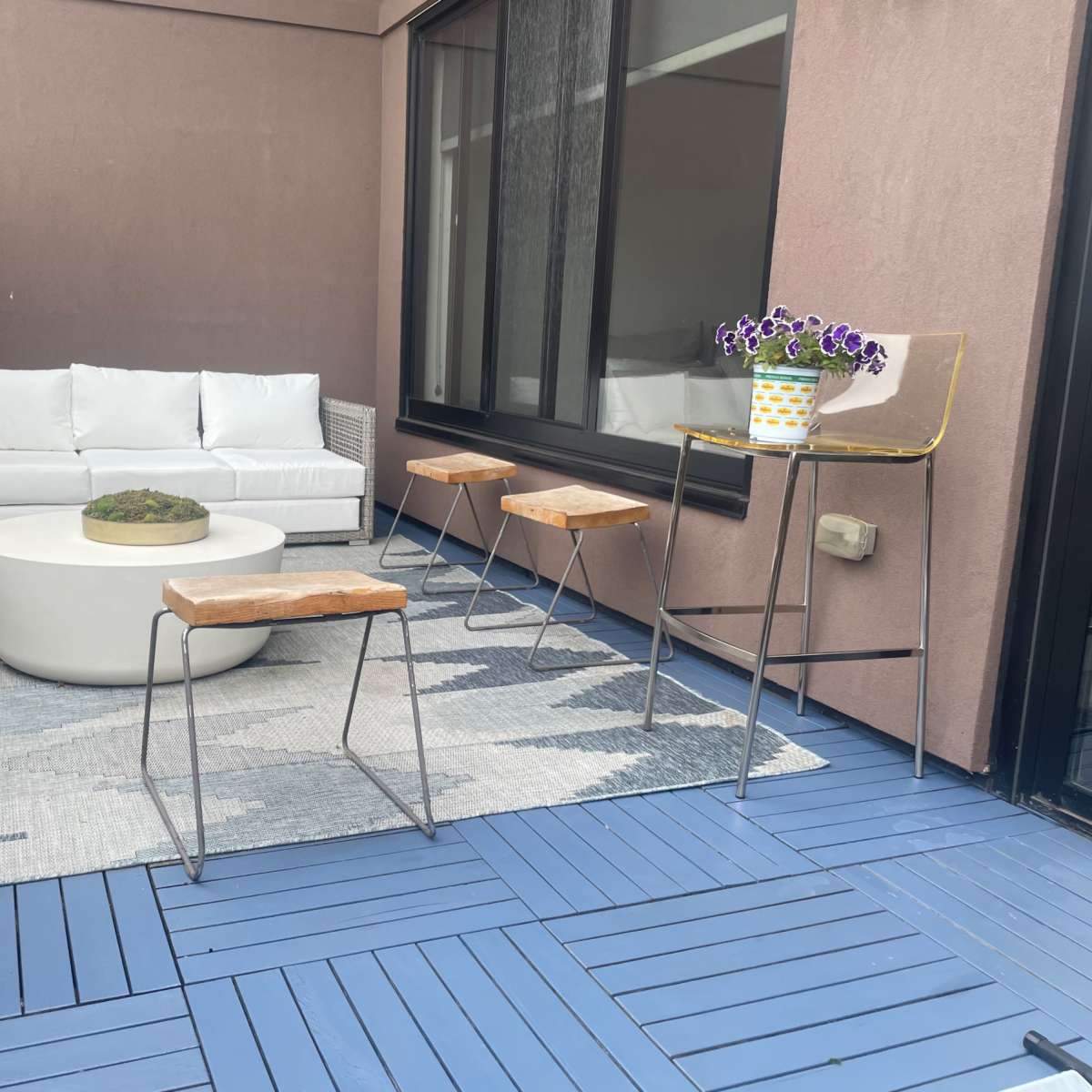 A modern balcony with a white sofa, circular coffee table, and several wooden stools arranged on a blue deck with a patterned rug.