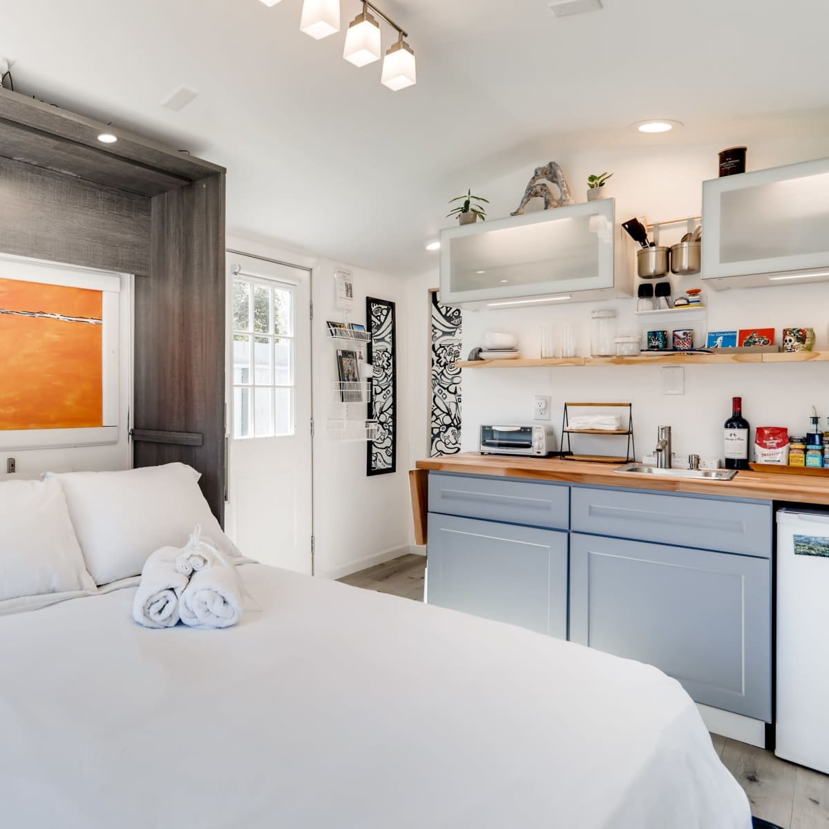 A small, modern studio apartment features a bed with white linens, a colorful artwork above it, and a kitchenette with wooden accents.