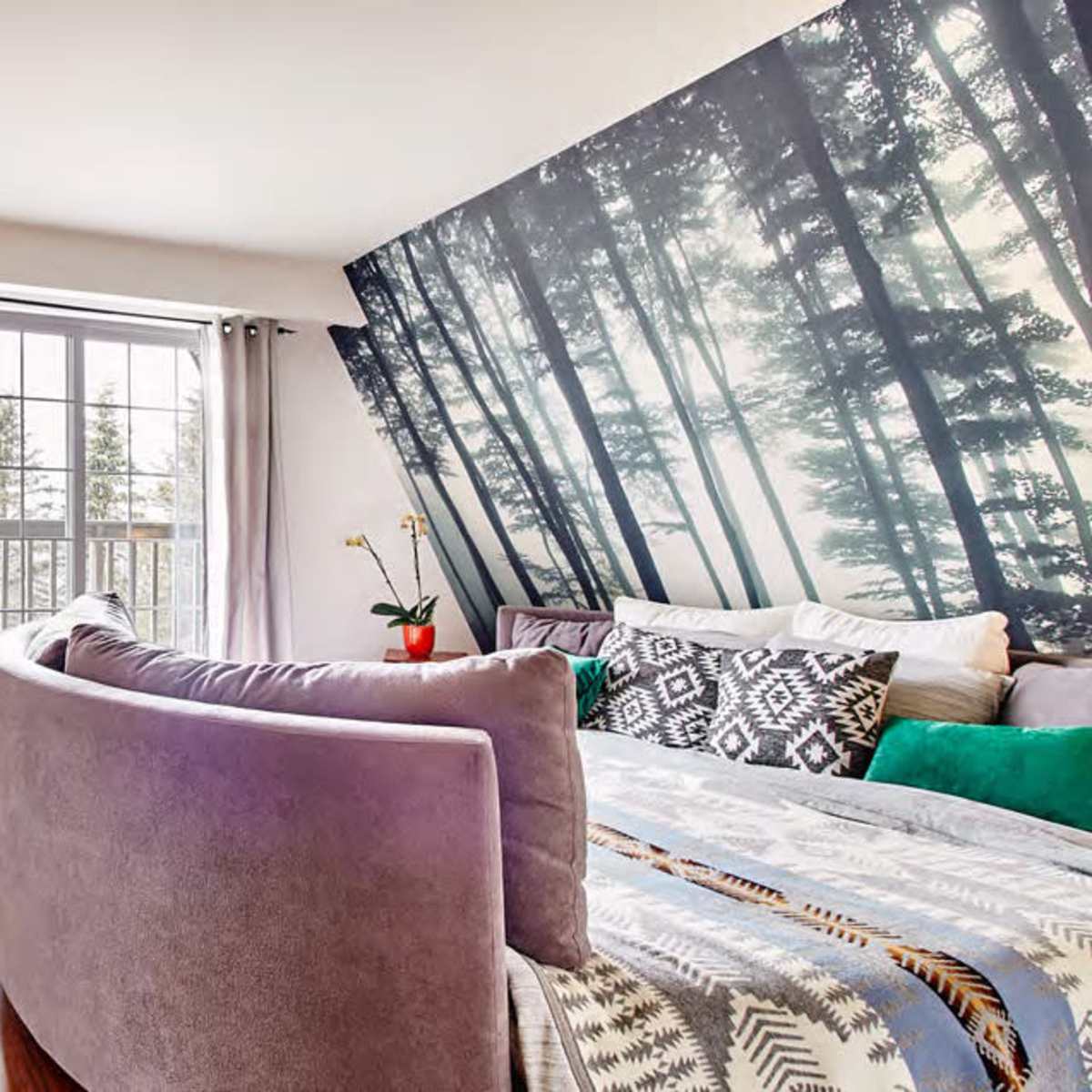 A modern bedroom features a curved sofa, a wooden desk with a lamp, and a large forest-themed mural on one wall.