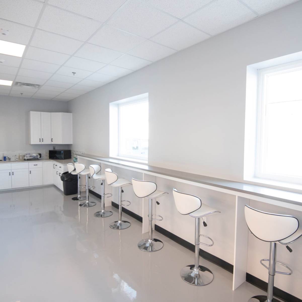 A modern kitchen with white cabinetry and high stools along a counter near two windows.
