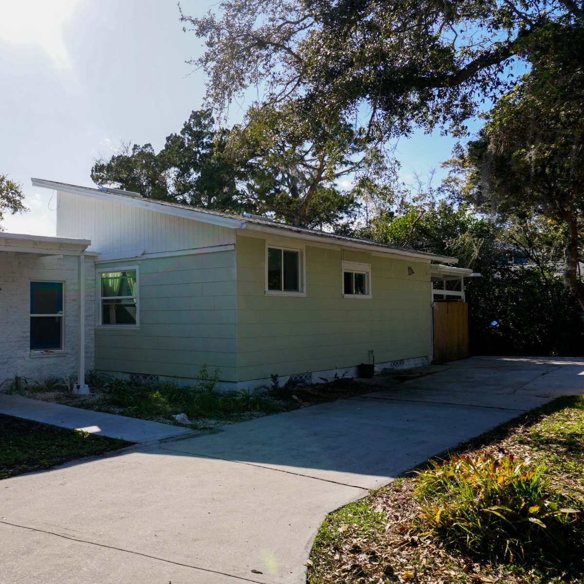 A modern single-story house with a mix of light green siding and a white exterior, accompanied by a concrete driveway and surrounding greenery.