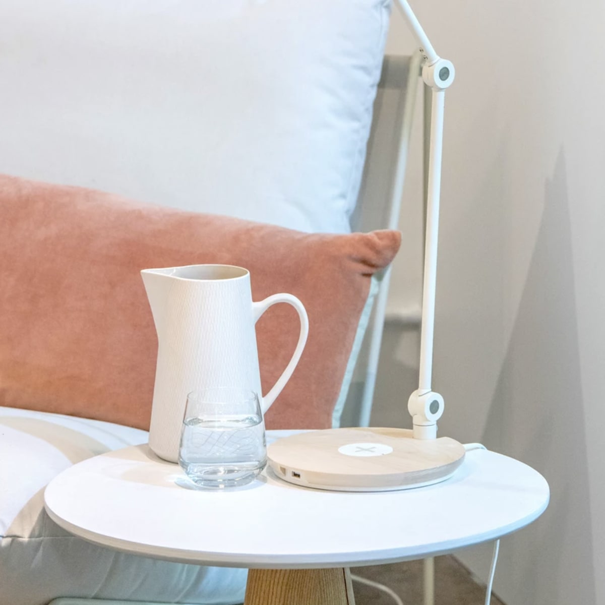 A white desk lamp illuminates a glass of water and a ceramic pitcher on a round side table beside a light-colored armchair with a pink cushion.