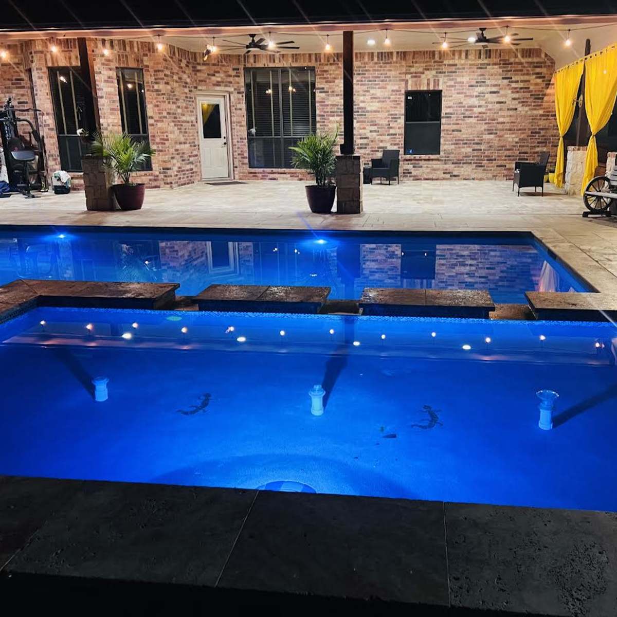 A well-lit outdoor pool area with a series of steps leading into the water and a brick house in the background.