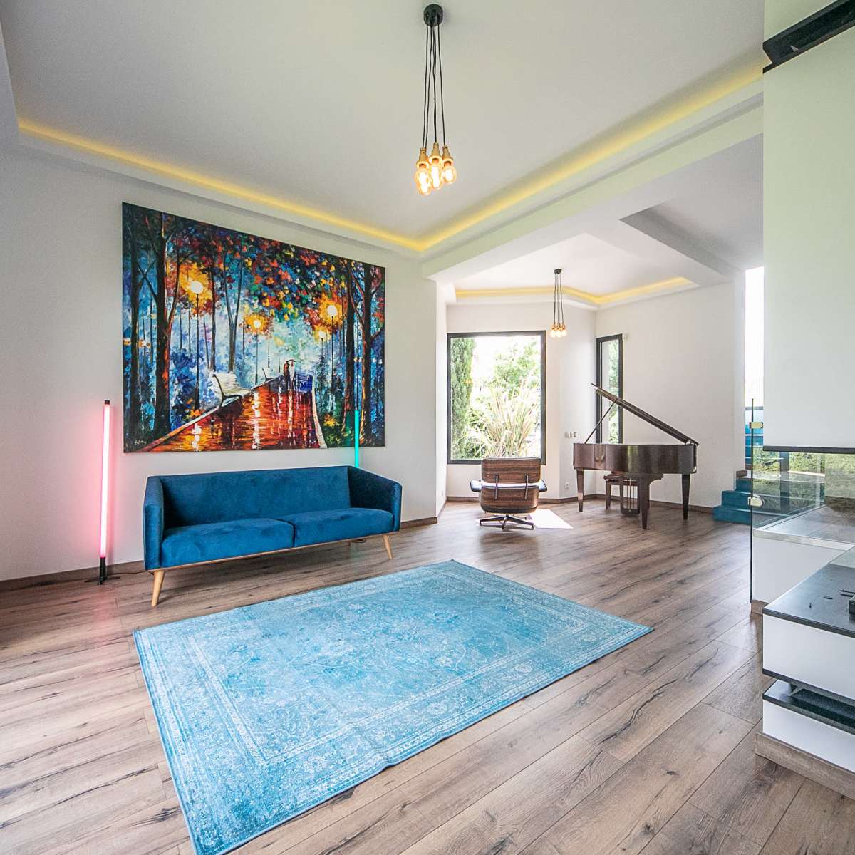 A living room features a blue couch, a colorful painting on the wall, a grand piano, and large windows that let in natural light.