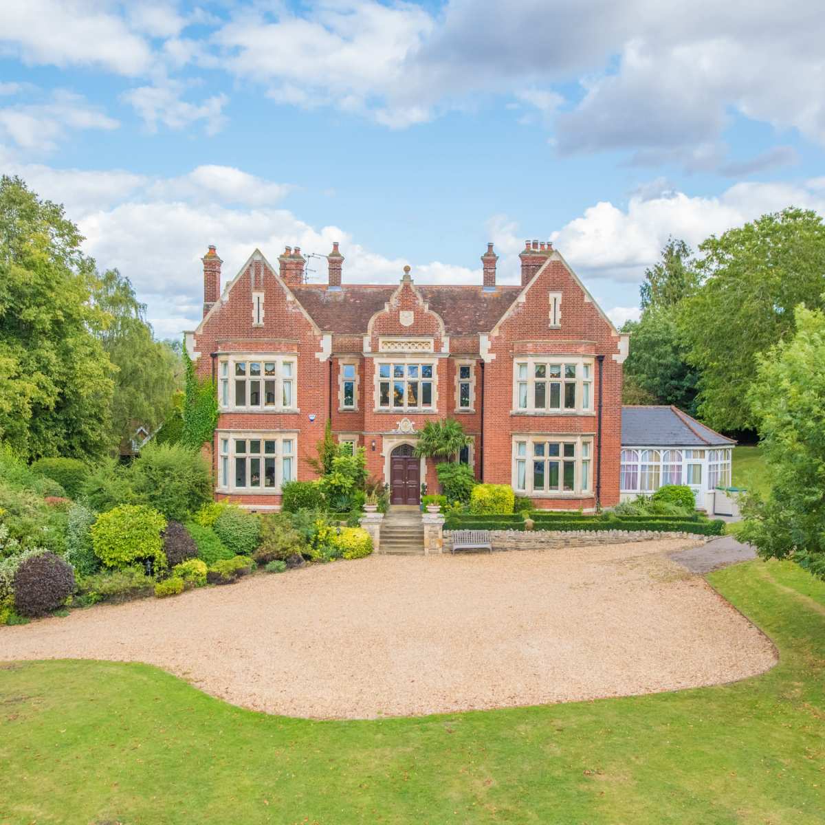 A large red brick mansion is surrounded by well-maintained gardens and trees, set against a partly cloudy sky.