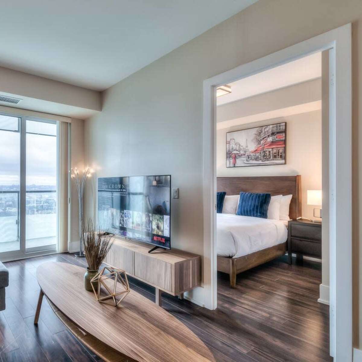 A modern living room features a blue sofa, a wooden coffee table, and a large TV, with sliding glass doors providing a view of the outside.