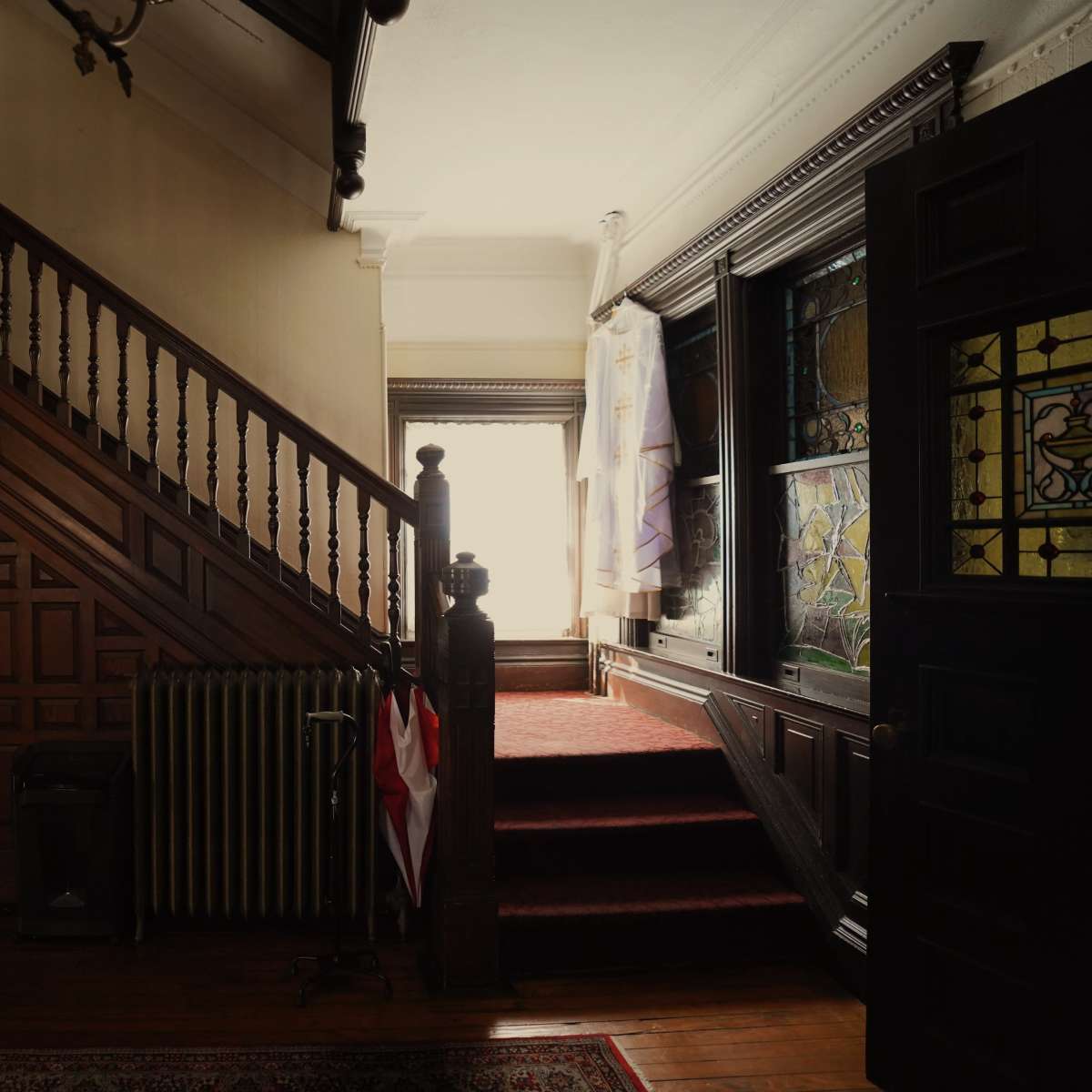 The entrance of a Victorian-style home with a wooden staircase leading to a brightly lit upper level, featuring ornate banisters and a stained glass window.