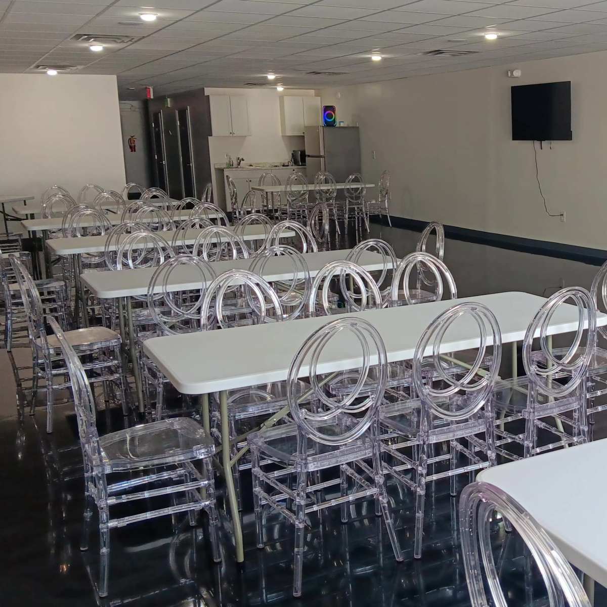 A spacious room with several white tables and transparent chairs arranged in neat rows, featuring a kitchenette and a wall-mounted television in the background.