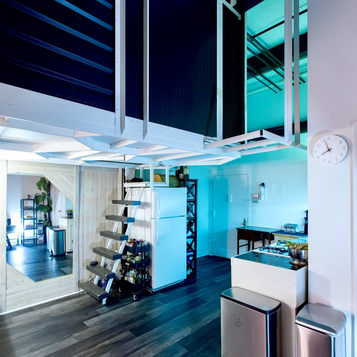 A modern, open-space interior with a staircase leading to a lofted area, a kitchen featuring appliances, and a large mirror reflecting the space.