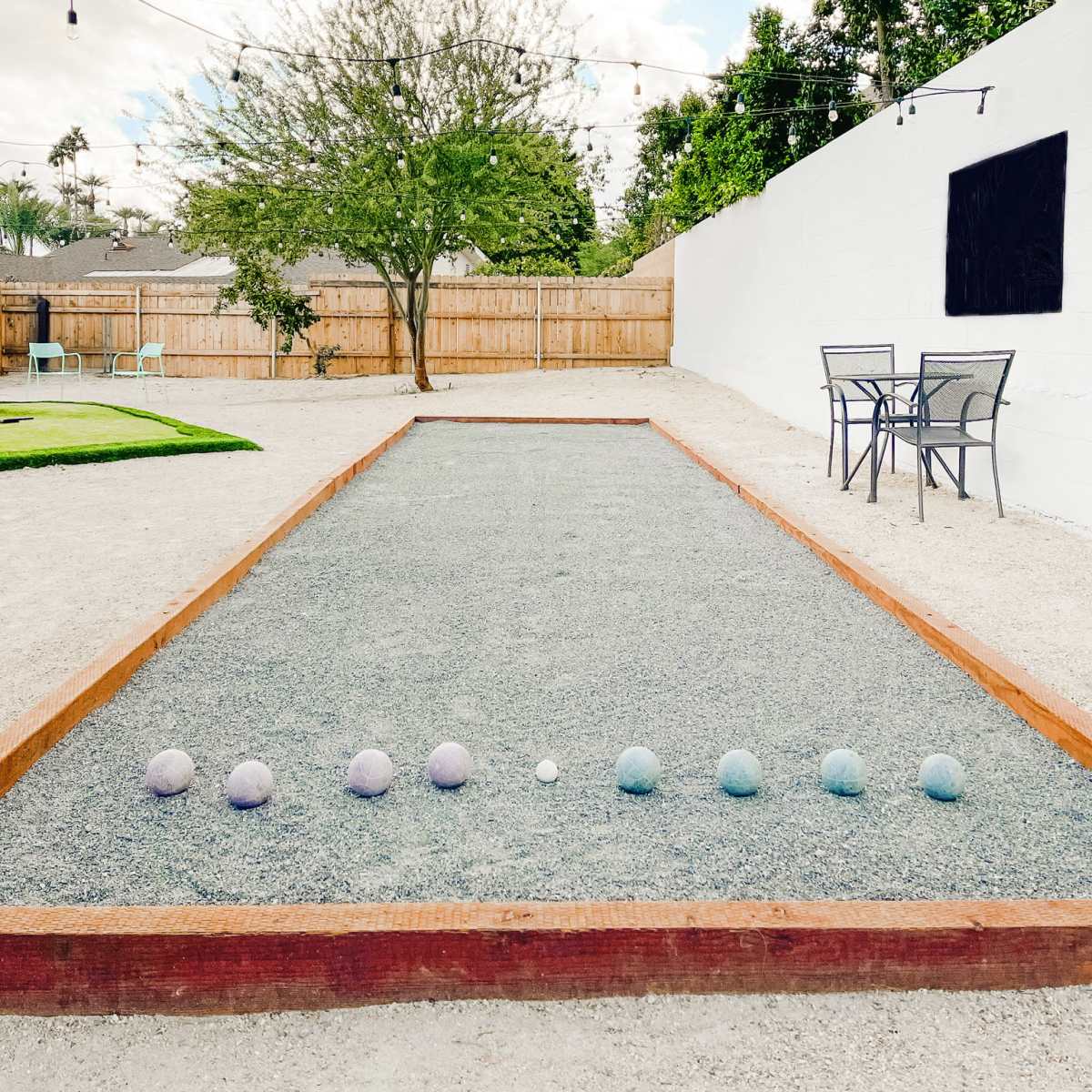 A gravel bocce court with colorful balls lined up, surrounded by greenery and string lights, is set against a white wall and a wooden fence.