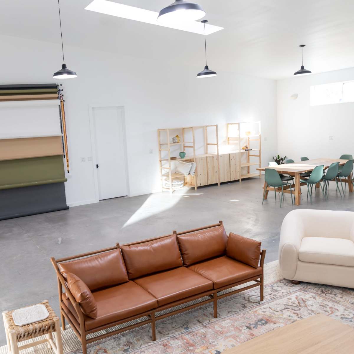 The interior of a spacious, modern room features a leather sofa, a light-colored armchair, a dining table with chairs, and a wall with a roll of fabric in various colors.