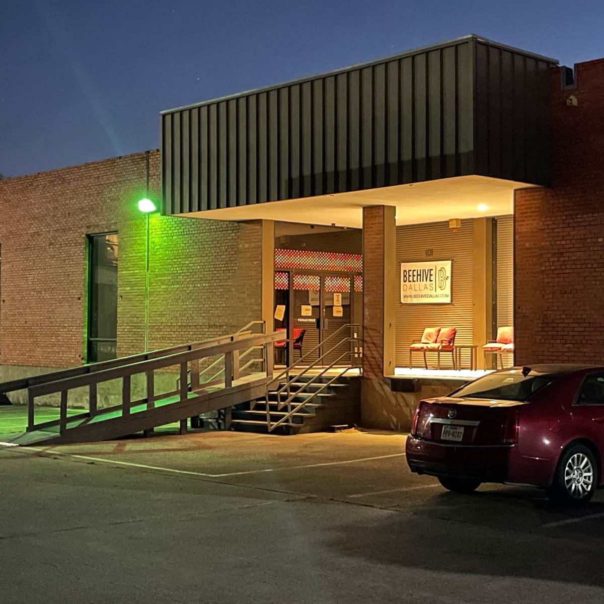 A building entrance with a ramp leading to a door, accompanied by a parked red car in a dimly lit parking lot.