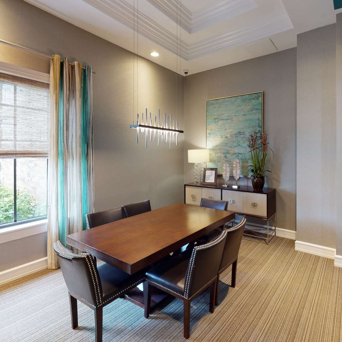 A well-lit office space featuring a wooden table surrounded by chairs, a console table with decor, and large windows with shades.