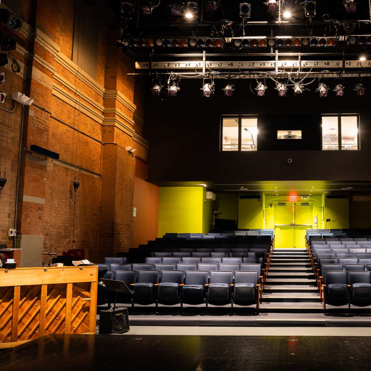 A theater interior with rows of blue seats facing a stage, illuminated by overhead lighting and featuring large windows above the entrance.