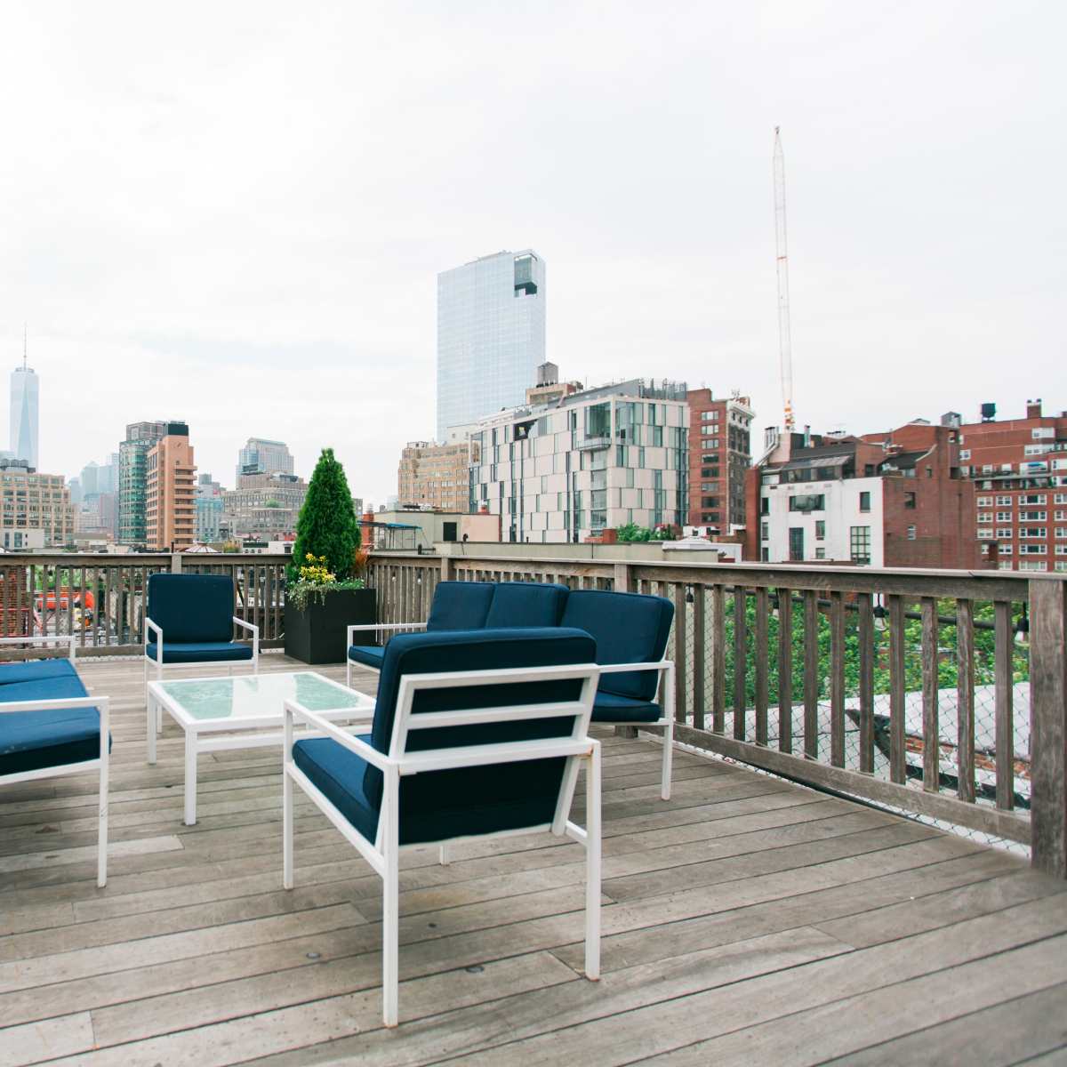 A rooftop deck features four blue chairs and a glass table, overlooking a city skyline with various buildings.