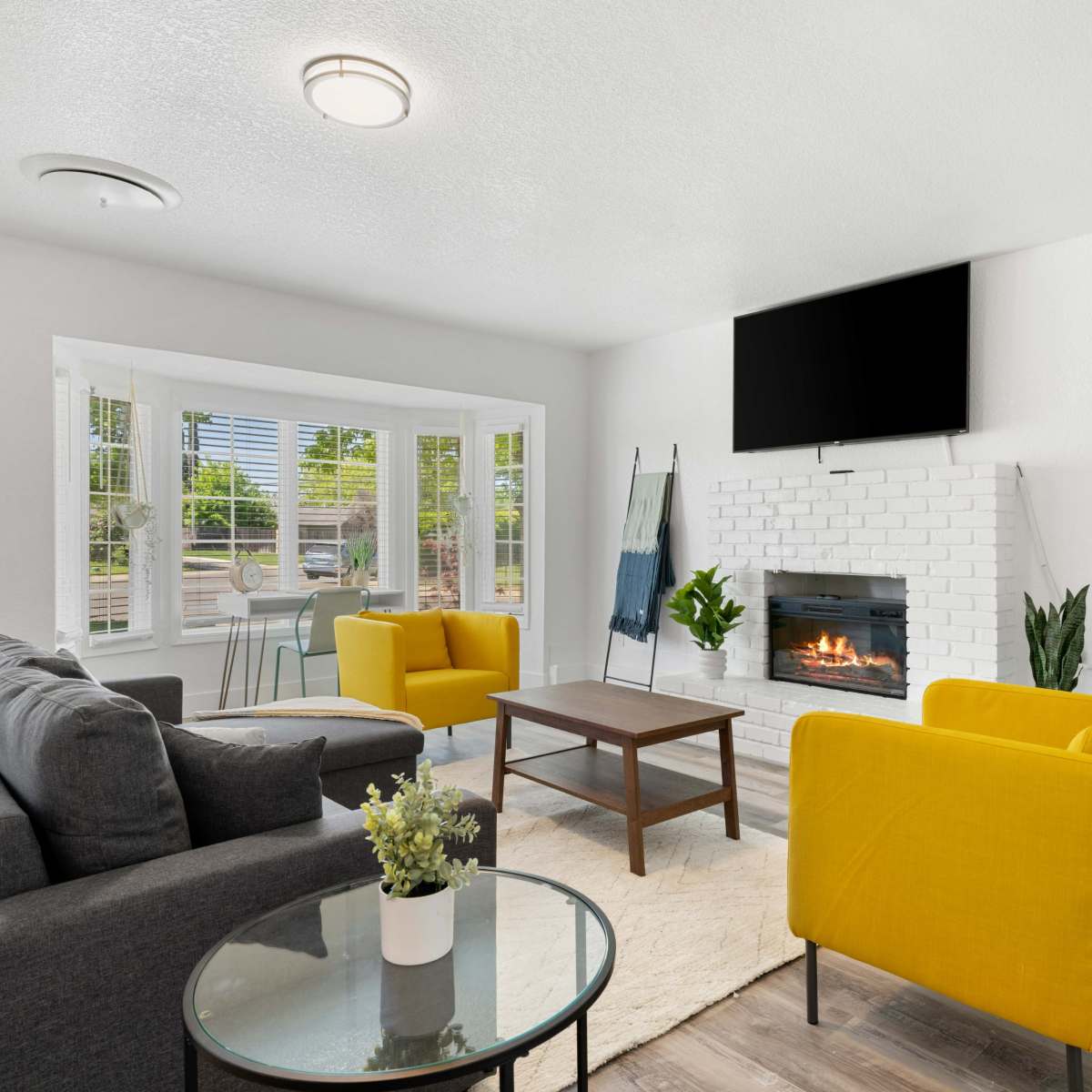 A modern living room features a gray sofa, two yellow armchairs, a coffee table, and a white brick fireplace beneath a wall-mounted TV.