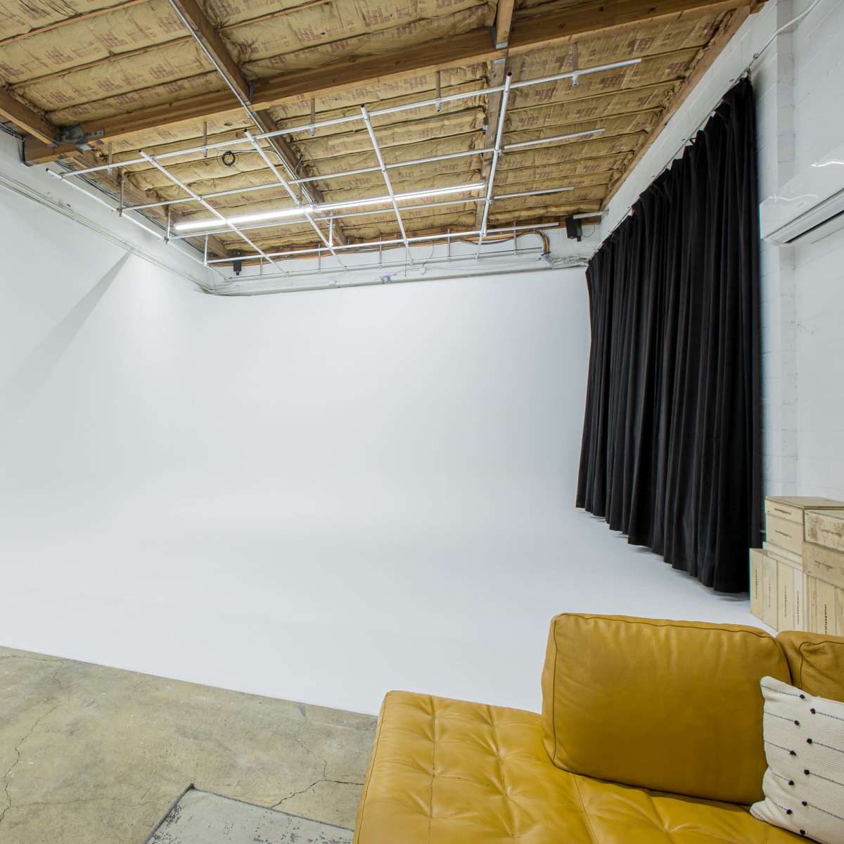 A spacious photography studio featuring a white backdrop, a yellow couch, a wooden desk with a chair, and a black curtain.