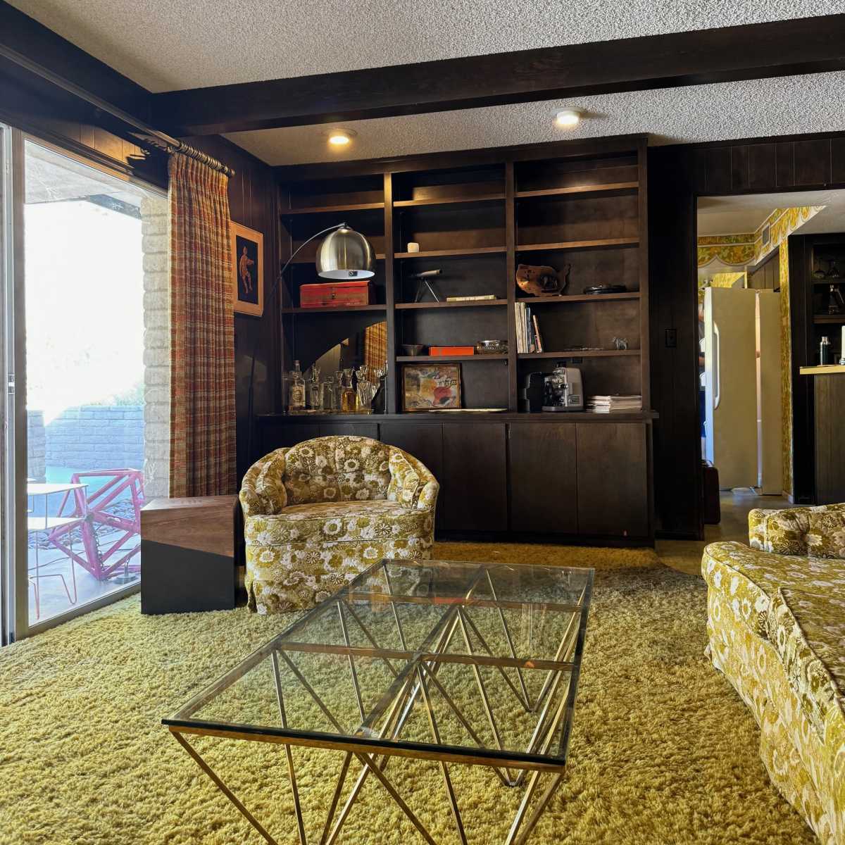 A mid-century modern living room with patterned armchairs, a glass coffee table, a dark wooden bookshelf, and sliding glass doors leading to an outdoor area.