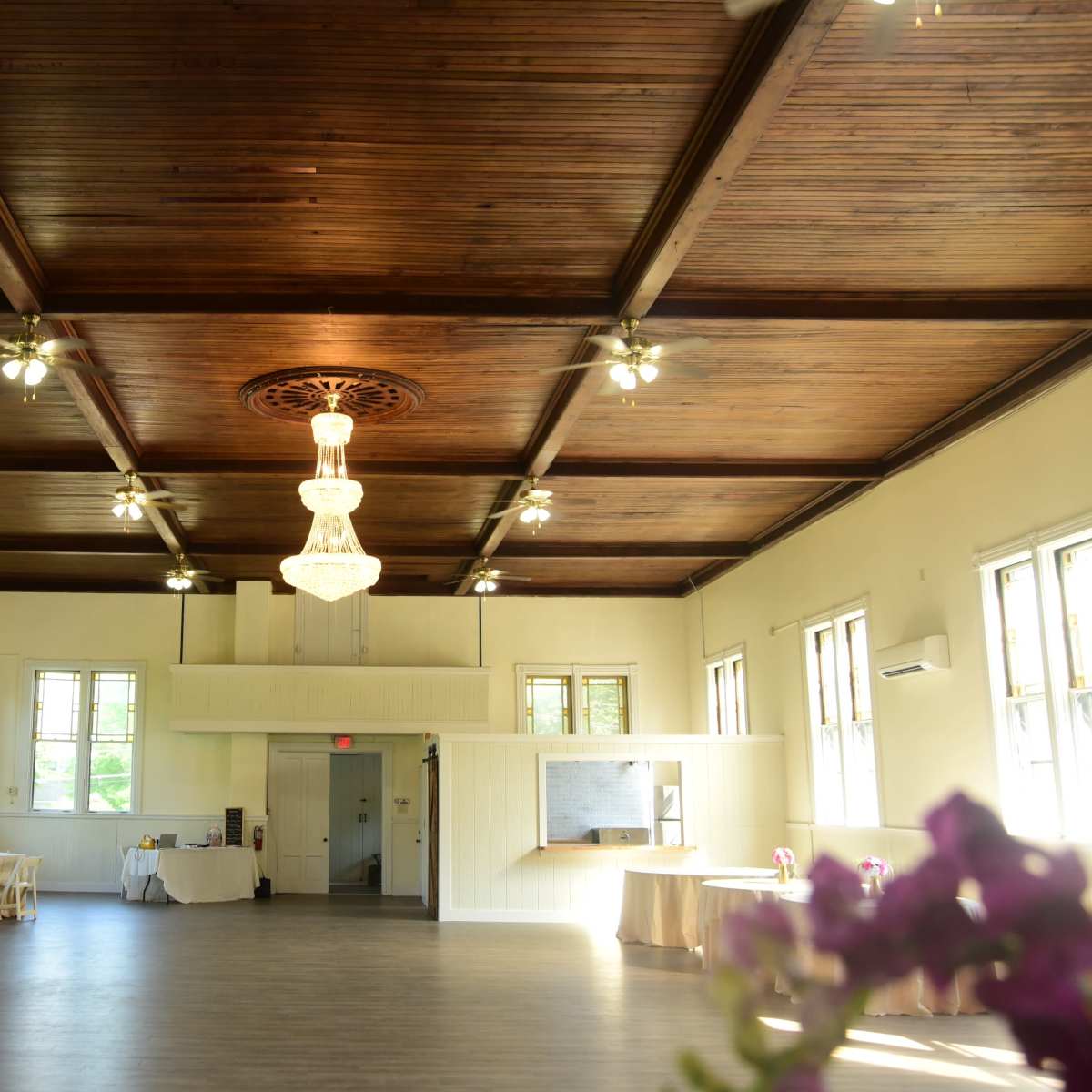 A spacious event hall with a wooden ceiling, a chandelier, and several tables arranged around the perimeter.