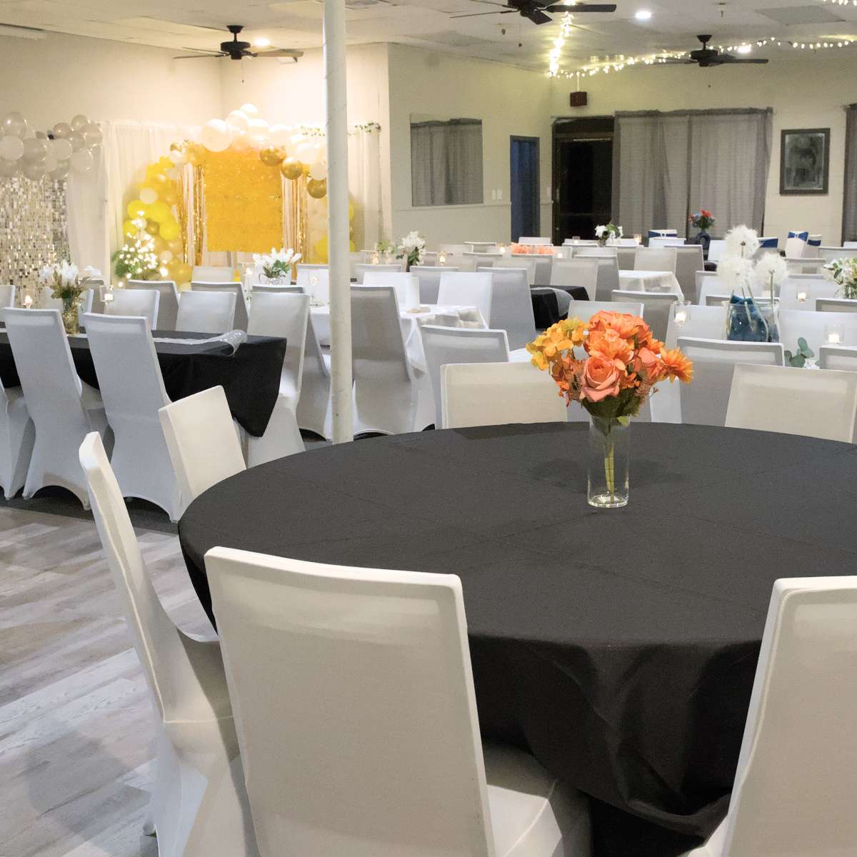 A decorated event space with round tables covered in black and white tablecloths, floral centerpieces, and a backdrop adorned with balloons.