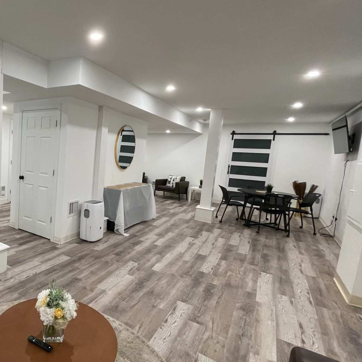 A well-lit, modern basement living space features a sitting area, dining table, and a kitchenette with a large mirror on the wall.