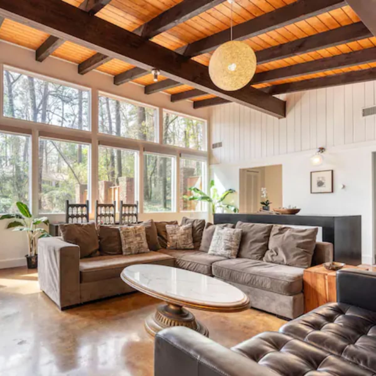 The living room features large windows, exposed wooden beams, a comfortable sectional sofa, and a central coffee table on a polished floor.
