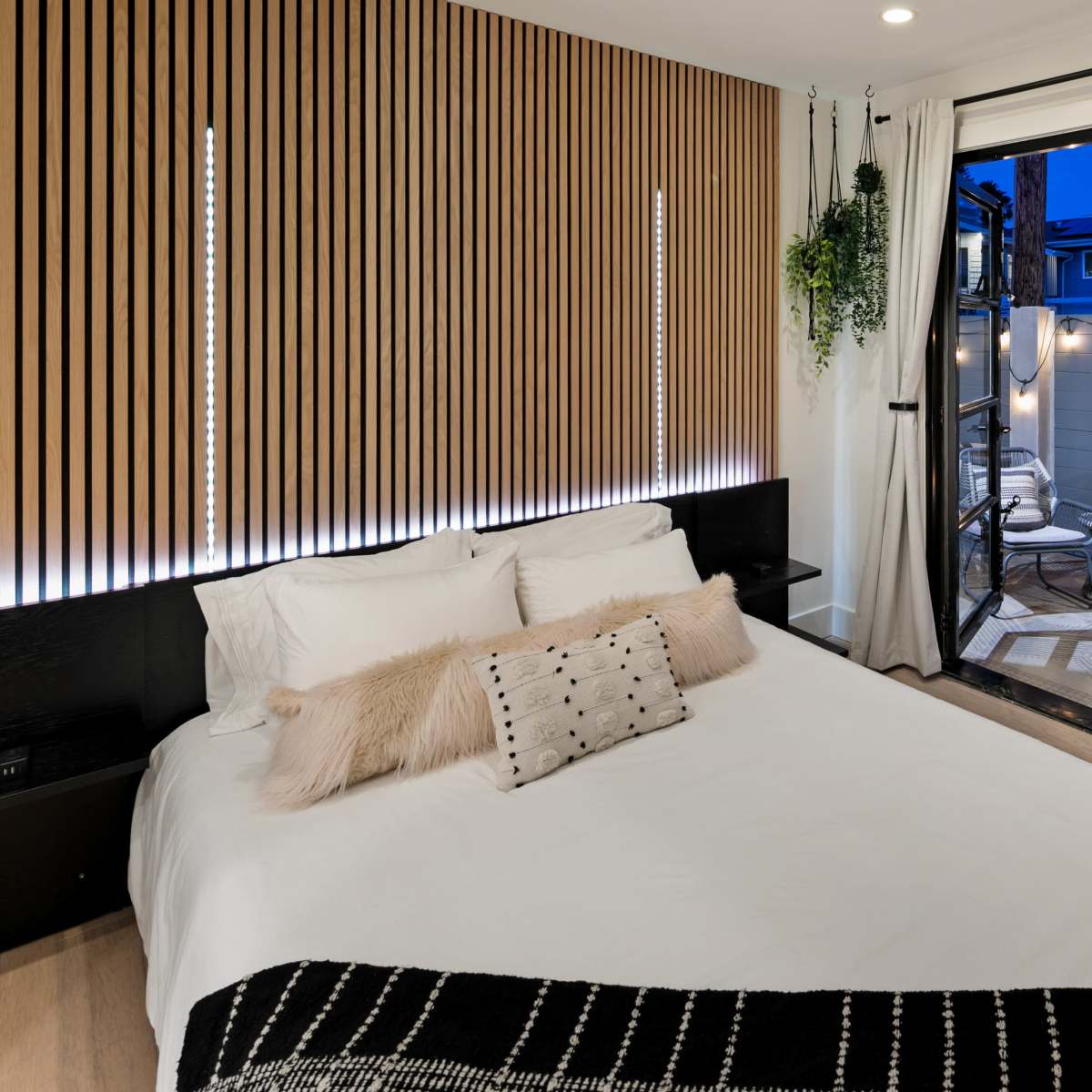 A modern bedroom features a bed with a decorative pillow, wooden paneling on the wall, and an open sliding door leading to a cozy outdoor seating area with a fire pit.