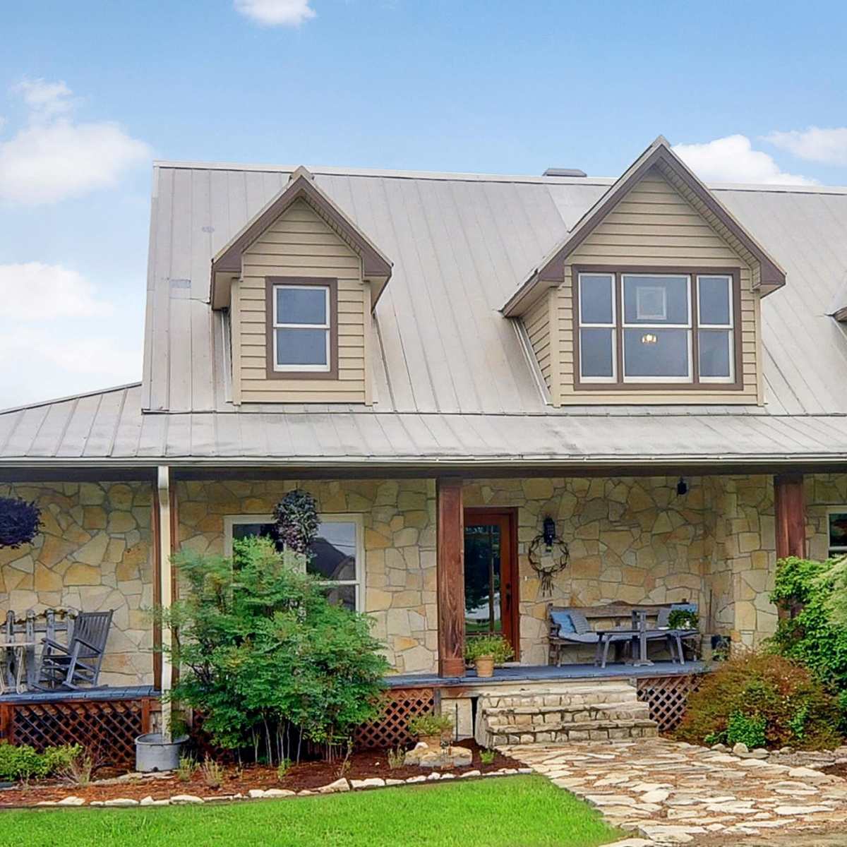 A two-story house with a stone facade, a metal roof, and a welcoming porch surrounded by greenery and landscaped grounds.