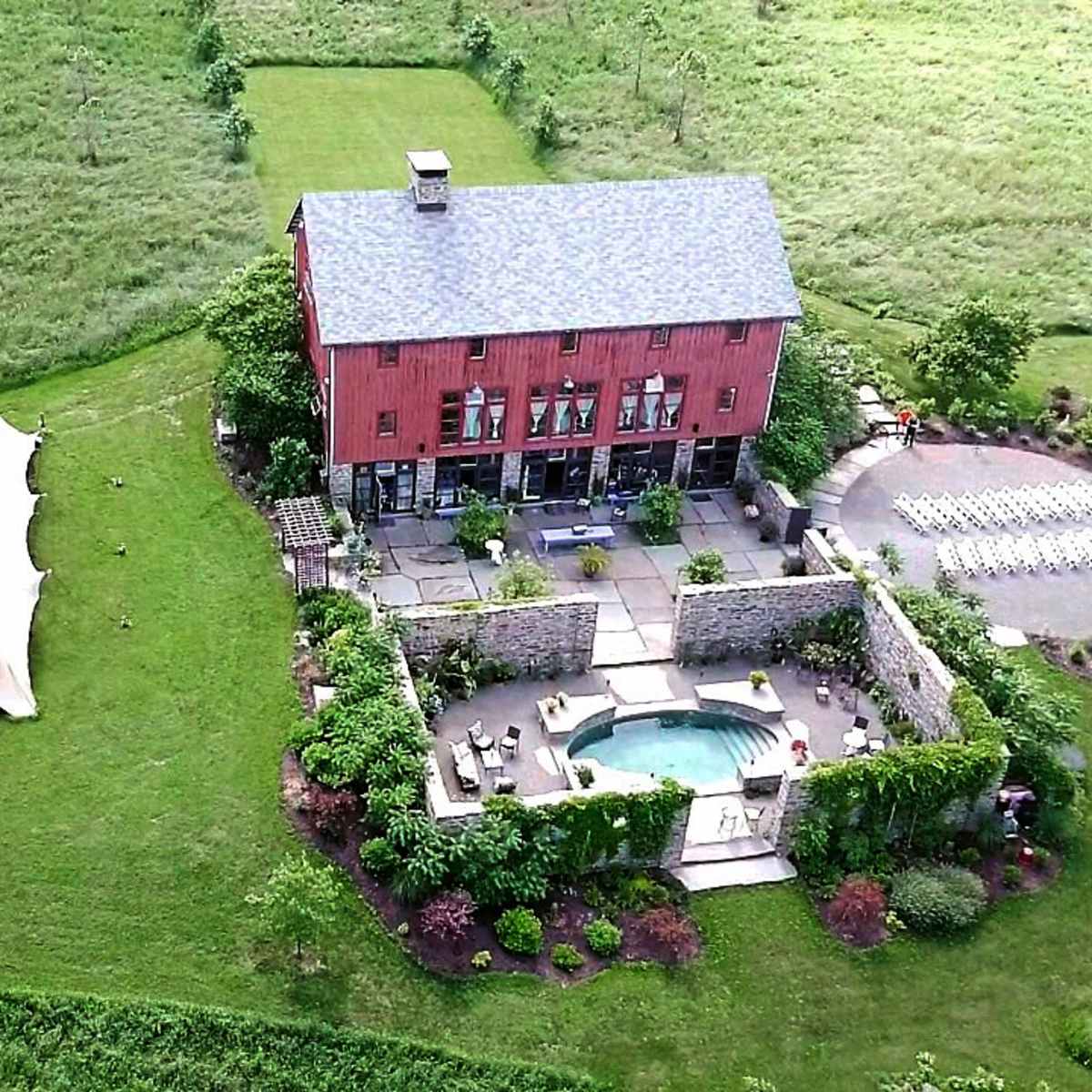 An aerial view shows a large red barn house surrounded by landscaped gardens, a pool, and a tented area on a grassy field.