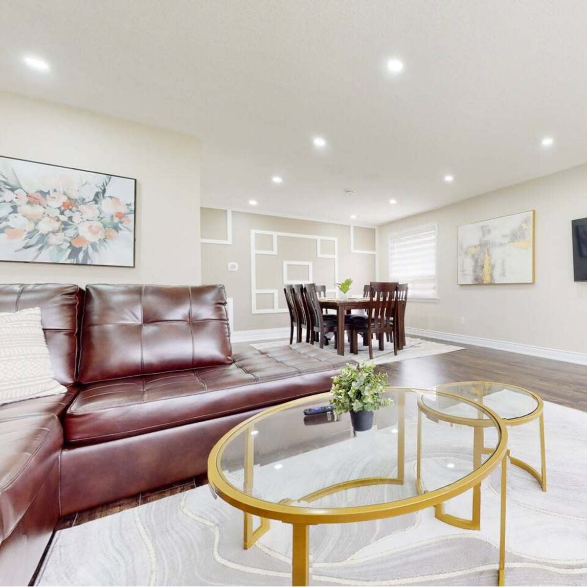 A modern living room features a brown leather sectional sofa, a glass coffee table, and a dining area with a wooden table and chairs, illuminated by ceiling lights.