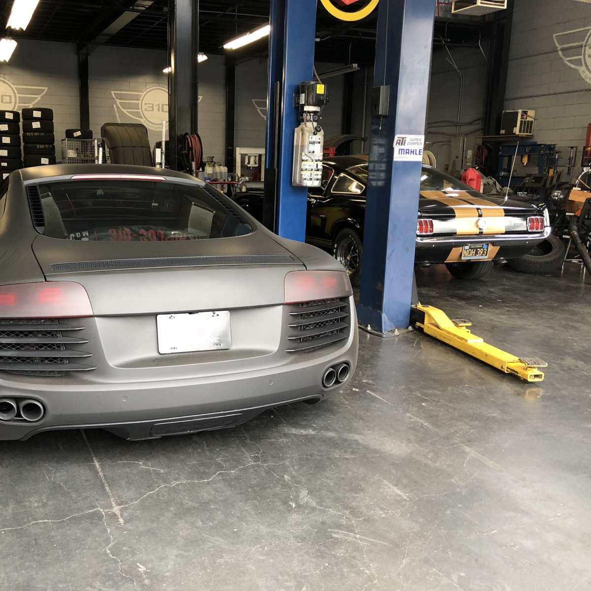 A matte black Audi R8 and a classic Mustang parked in a garage with car lifts and tools visible in the background.
