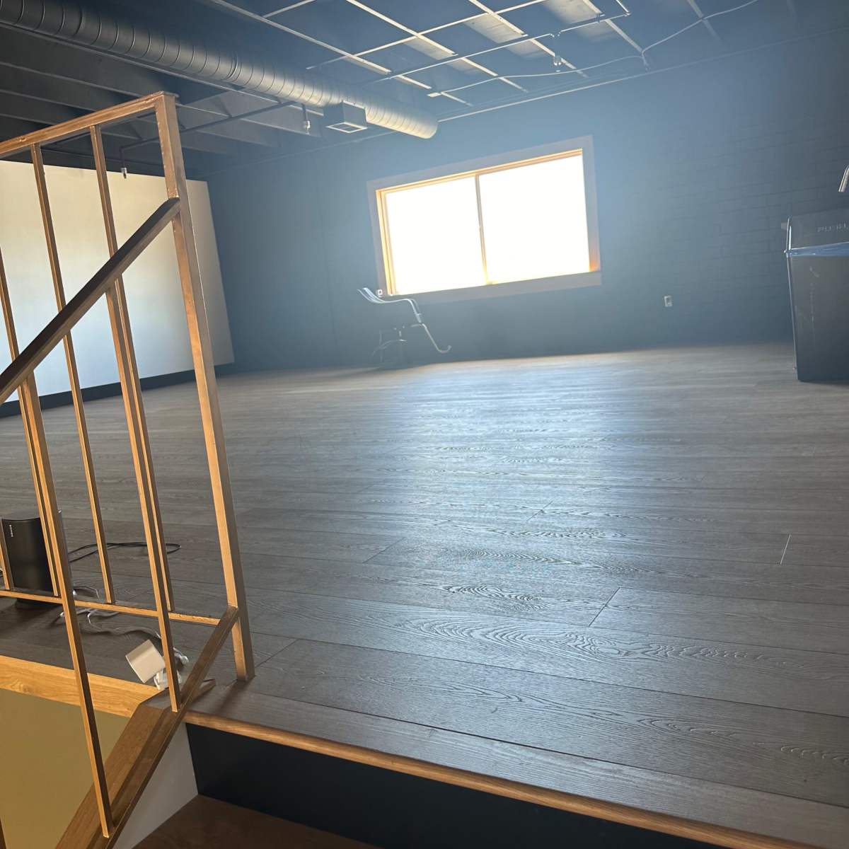 A spacious, empty room with wooden flooring and a large window allowing natural light, viewed from a staircase.
