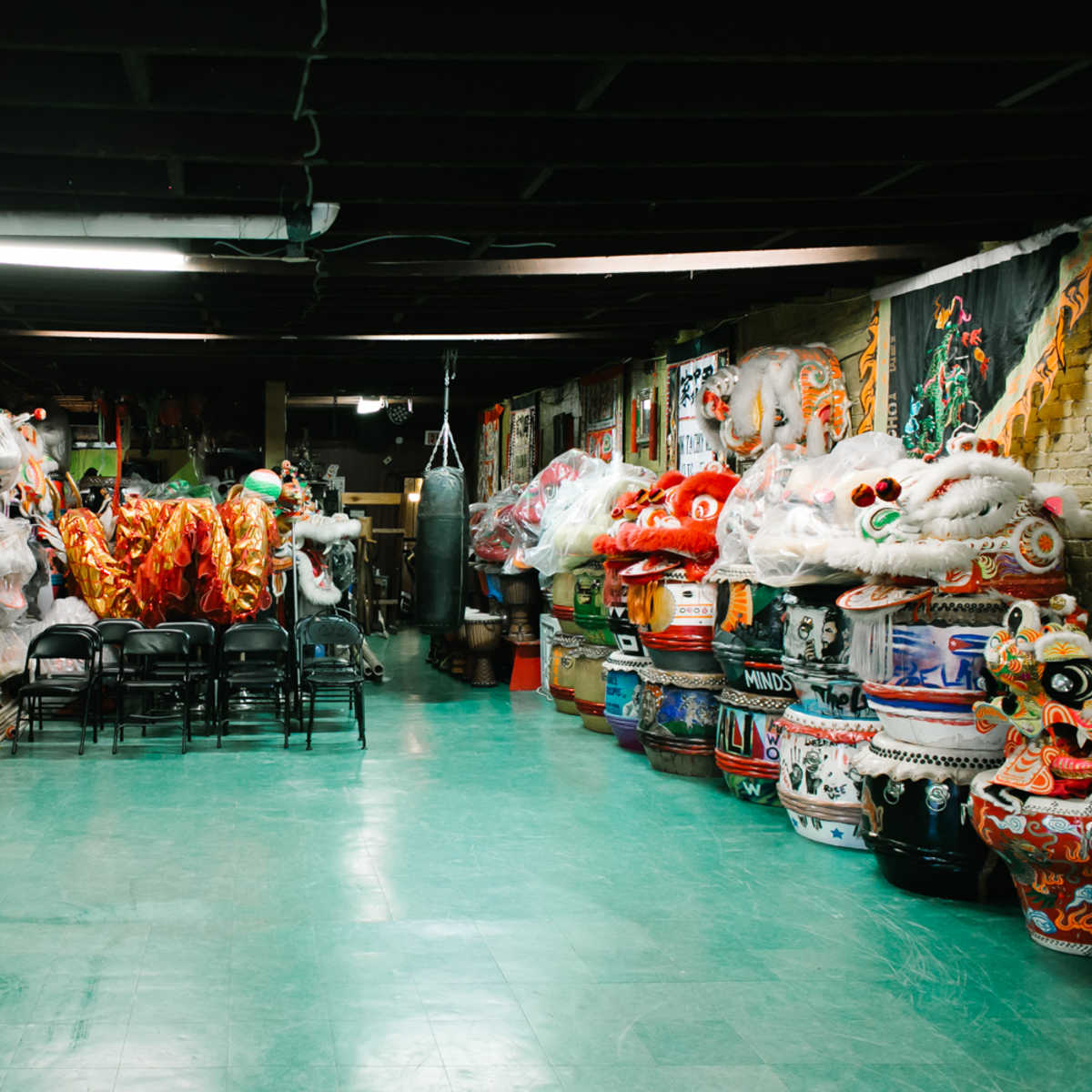 A dimly lit room filled with vibrant lion dance costumes and props, with black chairs arranged in the center.