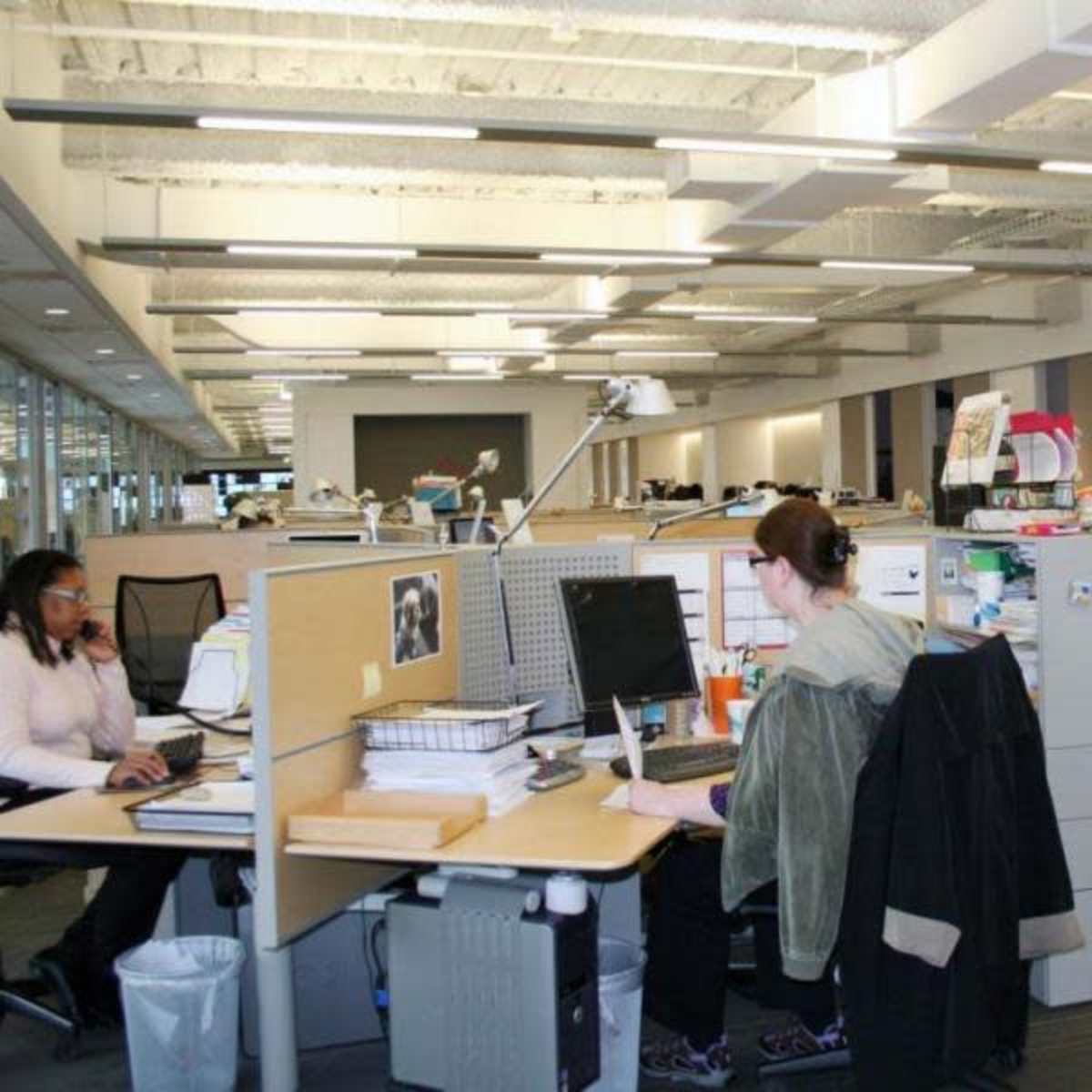 A modern office space with several workstations, each equipped with computers and office supplies, and individuals engaged in work activities.