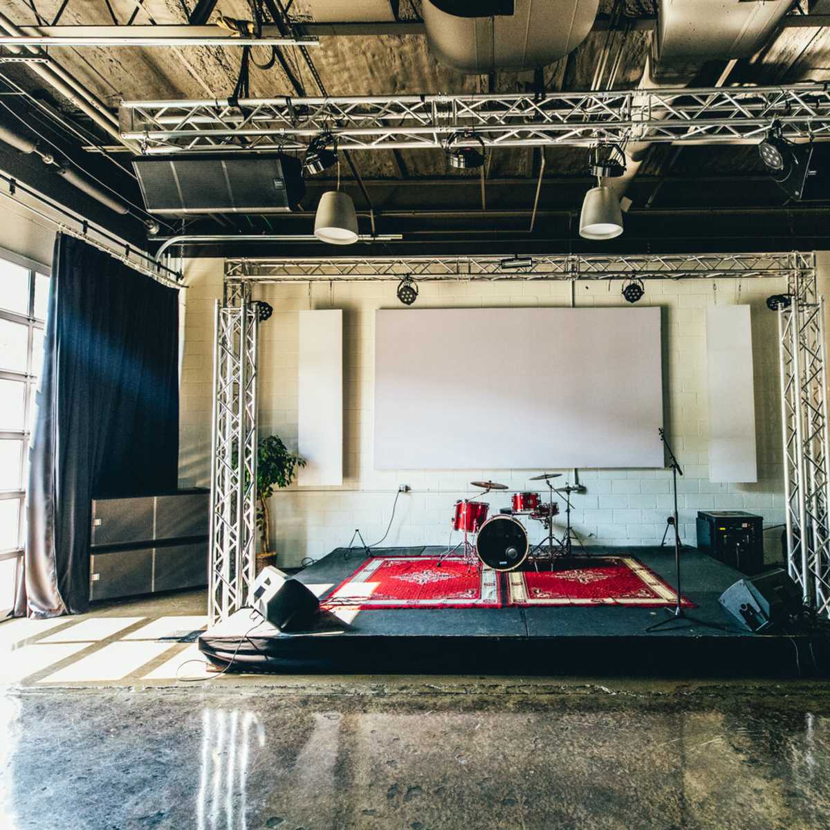 A stage set up for a performance, featuring a drum kit, amplifiers, and a large screen in a spacious venue with concrete flooring and large windows.