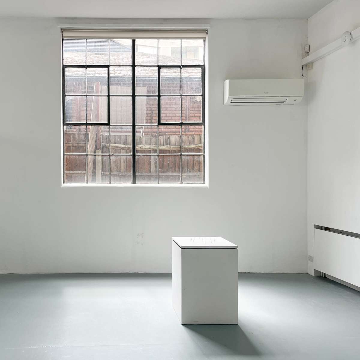 A minimalistic room with white walls, a single window letting in light, and a white pedestal in the center.
