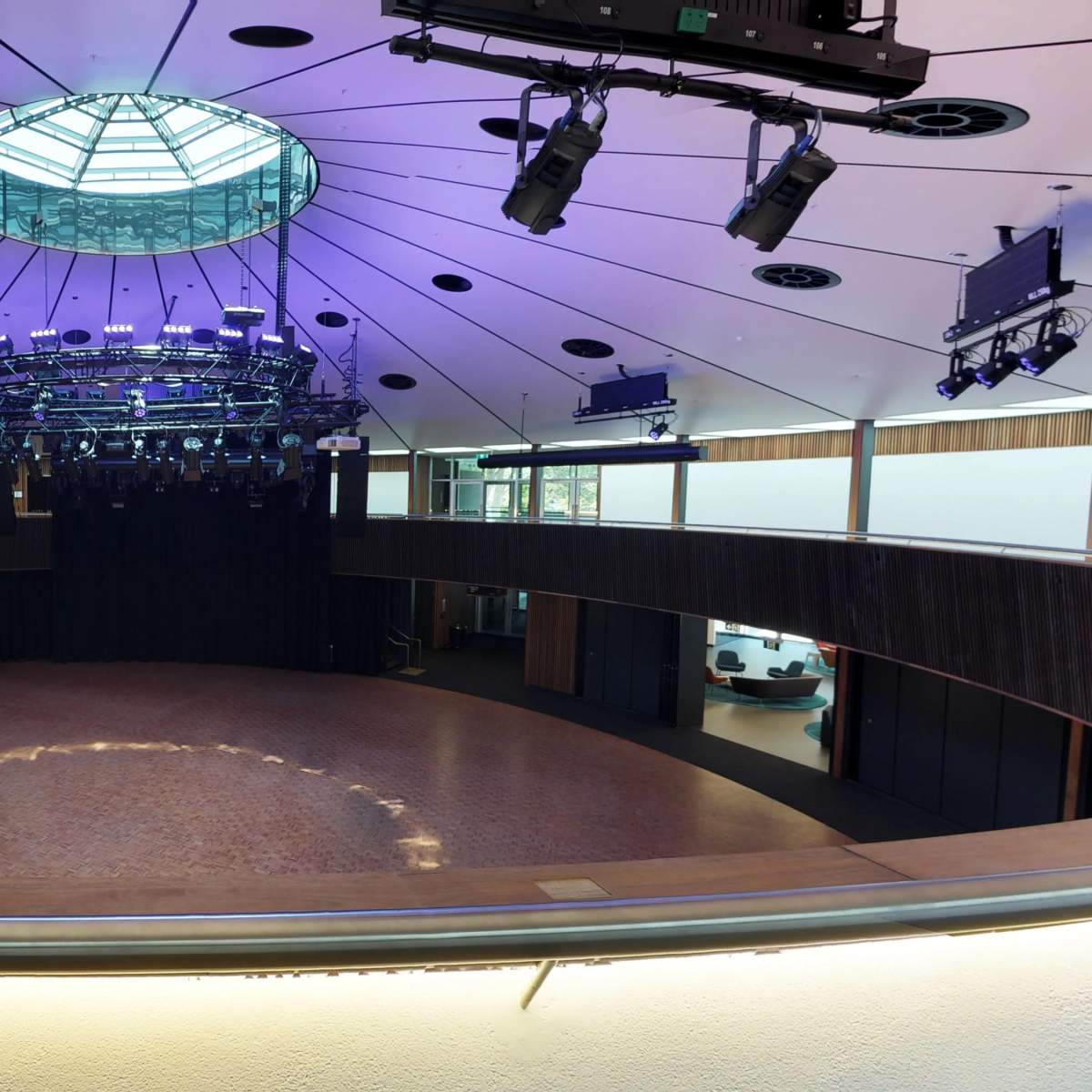 A modern auditorium with a circular stage, overhead lighting fixtures, and a glass dome ceiling, surrounded by seating areas.