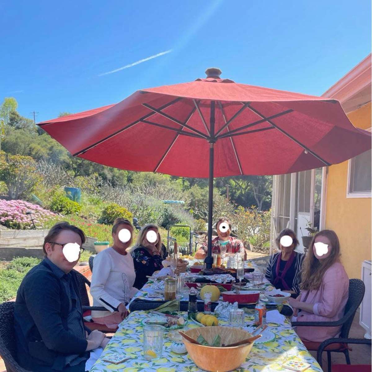 A group of six people sits around a table set with food and drinks under a large red umbrella in a sunny outdoor setting.