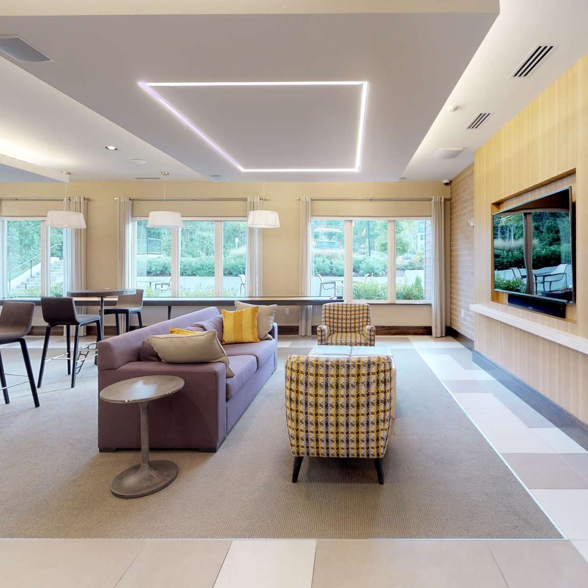 A modern lounge area with a purple sofa, a checkered armchair, and large windows letting in natural light.