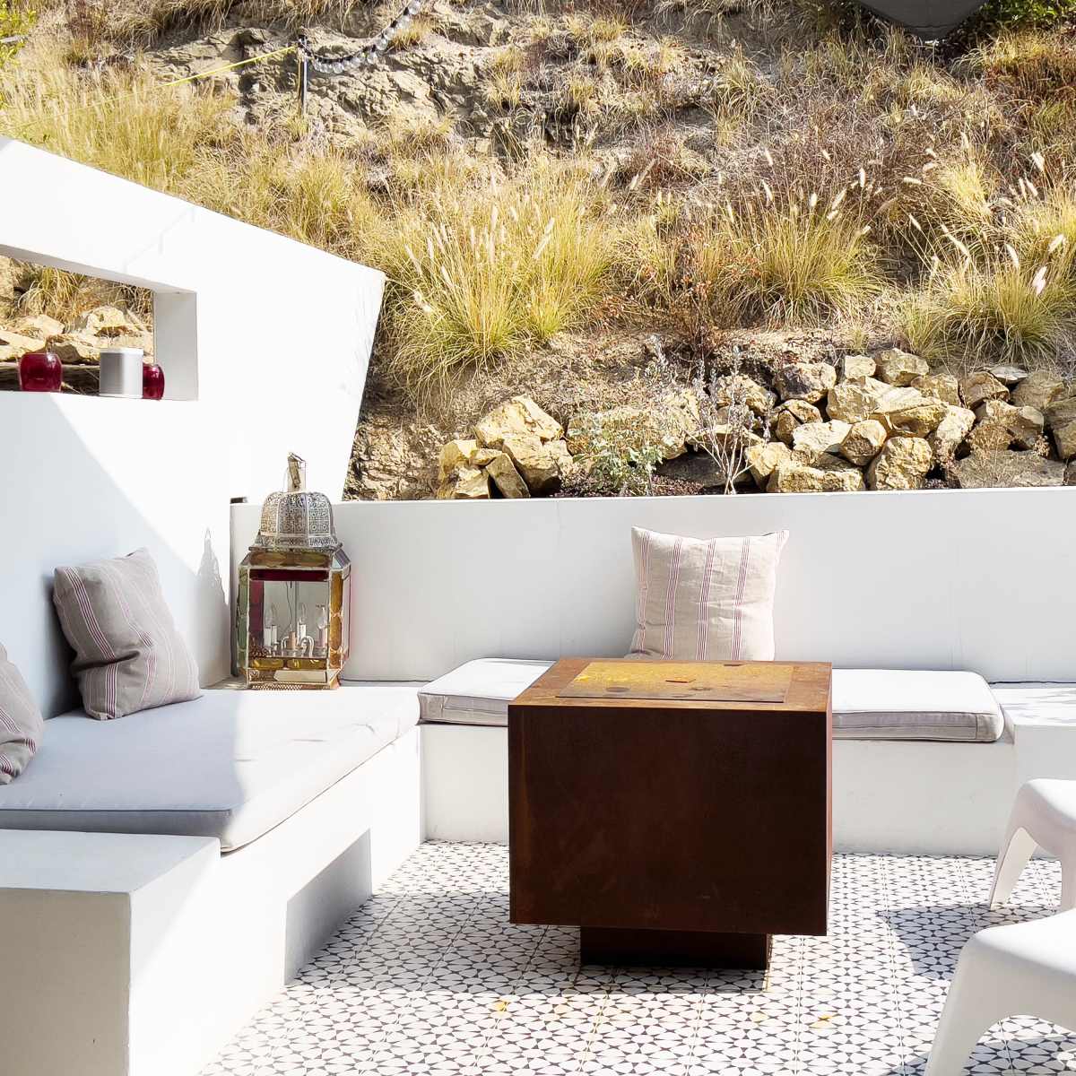 A modern outdoor seating area features a metal coffee table, surrounded by white couches and accented by decorative pillows, set against a backdrop of rocky terrain and dry grasses.